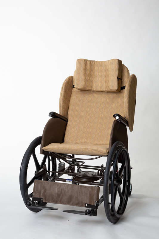 Picture of Rock N Go wheel chair fitted with lateral supports.  Cognac frame finish with padded cushion and standard head pillow in Tapestry fabric.  Fitted with standard footplates and heel/leg strap.
